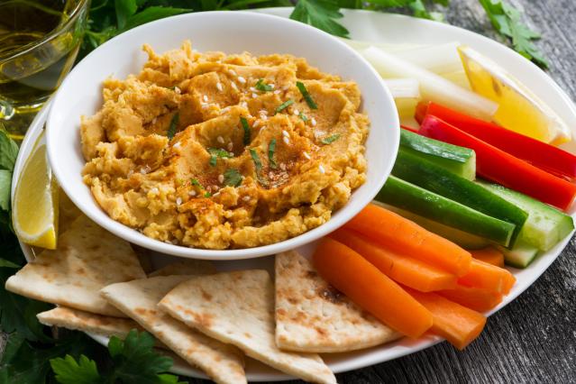 bowl of carrot and cumin spread garnished with herbs and sesame seeds, served with carrot sticks, cucumber sticks, capsicum strips, pita bread, and a lemon wedge on a plate.