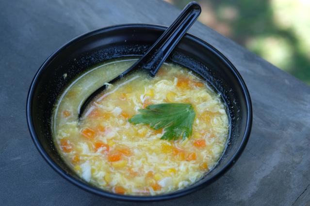 A black bowl of chicken and corn soup with visible corn kernels and diced vegetables in a light broth. A black soup spoon rests in the bowl, and a parsley leaf sits on top as a garnish.