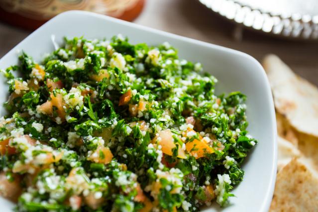 A white bowl filled with cauliflower tabbouleh made with finely chopped parsley, diced tomato, and cauliflower rice. Pieces of flatbread are blurred in the background. The salad is bright green and fresh-looking, with small flecks of tomato throughout.