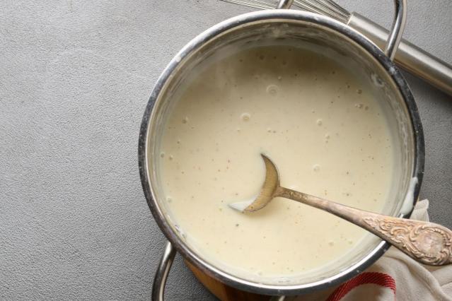 A saucepan filled with smooth, pale cheese sauce sits on a bench. A decorative metal spoon rests in the sauce, and a whisk is visible behind the saucepan.