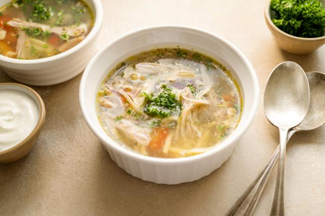 A white bowl of clear chicken soup containing shredded chicken, vegetables, herbs, and small pieces of stuffing in a light broth. Fresh parsley is sprinkled on top. Another bowl of soup, a small dish of yoghurt, and two spoons sit nearby on the table.