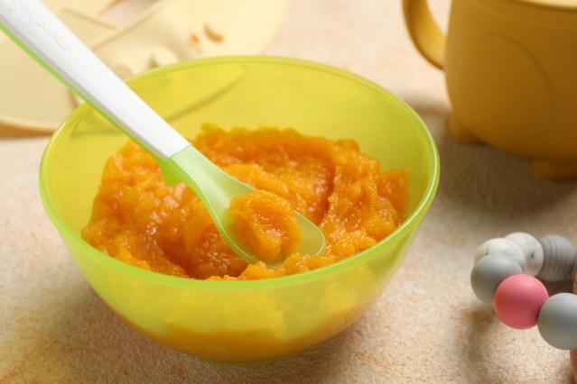 A green baby bowl filled with smooth orange chicken and carrot purée. A baby spoon rests in the purée with a spoonful lifted.