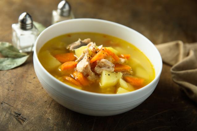 A white bowl of chicken and vegetable soup filled with pieces of chicken, sliced carrots, and diced potatoes in a clear golden broth. The bowl sits on a wooden table beside a folded cloth, with salt and pepper shakers blurred in the background.