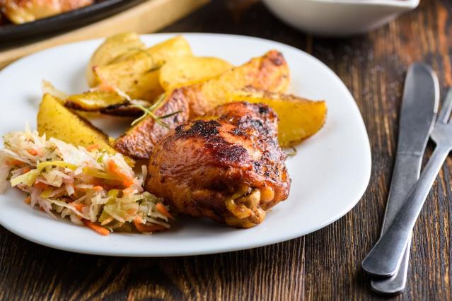A white plate holding a roasted chicken drumstick, golden potato wedges, and a serving of coleslaw made with shredded cabbage and carrot. The plate sits on a wooden table beside a knife and fork. Another dish is blurred in the background.