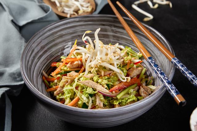 A bowl of chicken satay and crispy noodle salad made with shredded chicken, lettuce, carrot, cucumber, red onion, and bean sprouts. The salad is topped with extra bean sprouts and served with a pair of chopsticks resting on the side of the bowl.