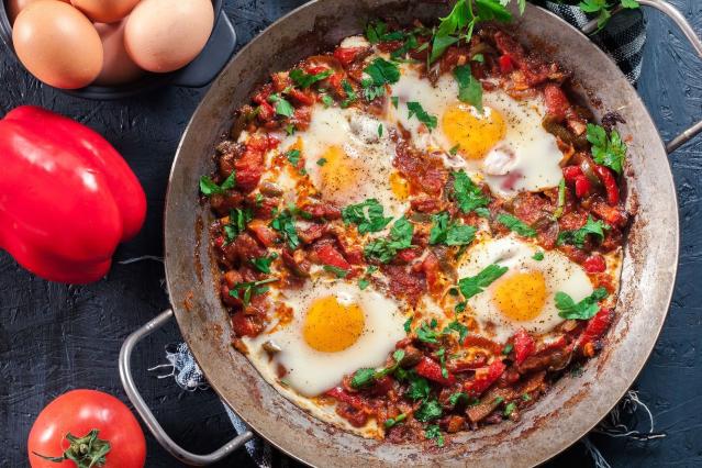 A frying pan filled with chilli beans and vegetables topped with four cooked eggs. The dish contains tomatoes, red capsicum, onion and herbs, with chopped parsley sprinkled over the top. Whole eggs, a red capsicum and a tomato sit beside the pan.