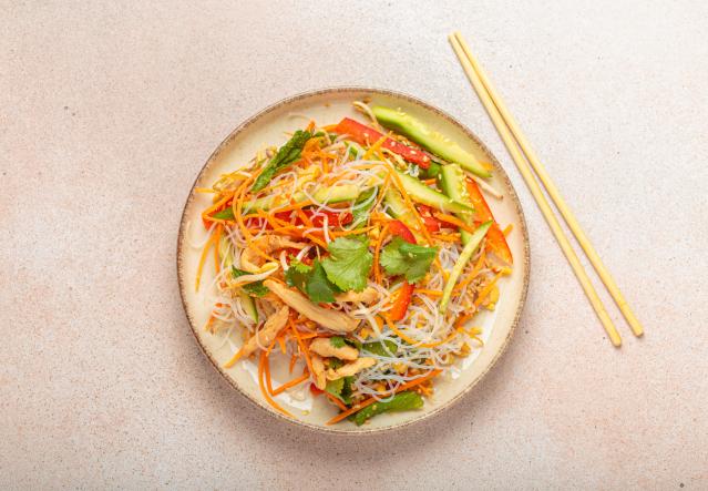 A plate of chilli chicken vermicelli salad viewed from above. The salad contains rice vermicelli noodles, shredded chicken, carrot, cucumber, red capsicum, bean sprouts, fresh herbs. A pair of chopsticks sits beside the plate.
