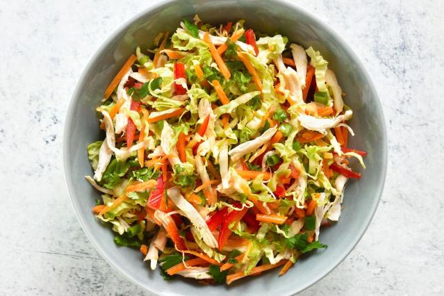 A bowl of coconut chicken slaw viewed from above. The slaw contains shredded chicken, finely sliced cabbage, carrot, red capsicum, spring onion and fresh herbs mixed together in a light dressing. The colourful salad is served in a grey bowl on a pale surface.