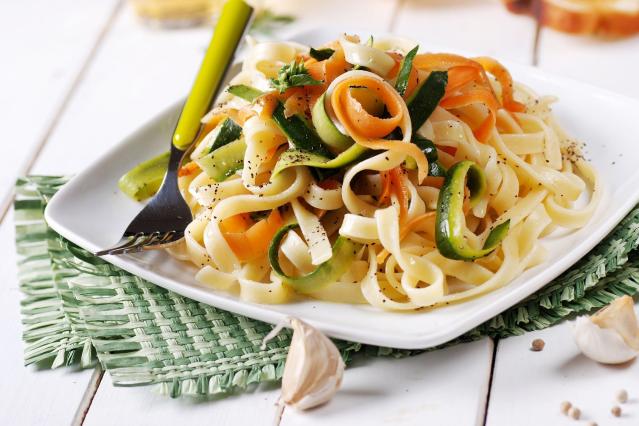 A plate of courgette pasta carbonara made with ribbon pasta, thin strips of courgette and carrot, and black pepper. The pasta is served on a white square plate with a fork, with garlic cloves and seasonings nearby.