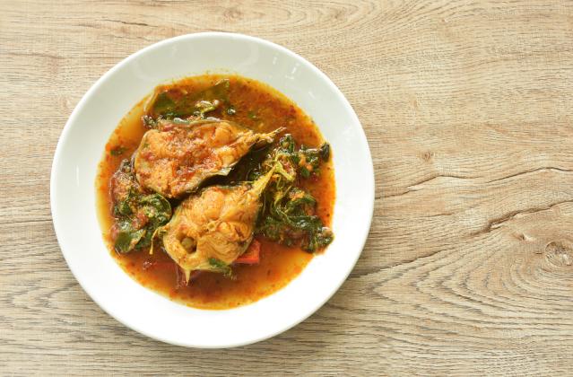 A bowl of eggplant curry with mackerel viewed from above. The curry contains pieces of mackerel and eggplant in a rich red-orange sauce with leafy greens and herbs. It is served in a white bowl on a light wooden table.