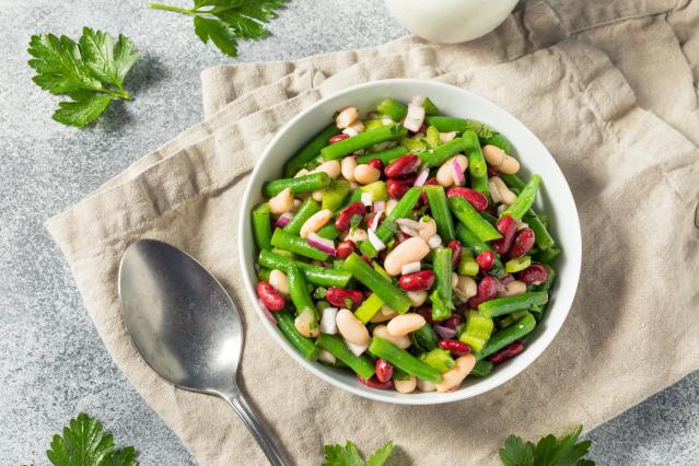 A bowl of green bean salad viewed from above. The salad contains green beans, red kidney beans, white beans, chopped red onion and celery, mixed with fresh herbs. The bowl sits on a beige cloth beside a large spoon, with parsley leaves scattered around.