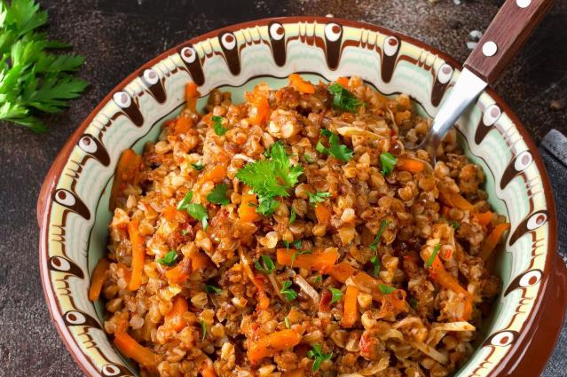 A decorative bowl filled with kai si ming made with pearl barley. The dish contains cooked barley mixed with shredded meat, carrot, onion, and seasonings, and is garnished with chopped parsley. A spoon rests in the bowl, which sits on a dark tabletop.