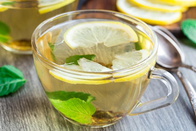 A clear glass cup of herbal tea containing floating lemon slices and green leaves. The cup sits on a wooden surface beside a spoon, with additional lemon slices and a second glass of tea blurred in the background. The tea is pale golden in colour and appears freshly prepared.