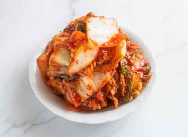 A white plate piled with kimchi, a traditional fermented cabbage dish. The cabbage leaves are coated in a bright red chilli seasoning, with visible layers of pale cabbage and green leafy pieces. The plate sits on a light-coloured surface and is shown against a plain background.