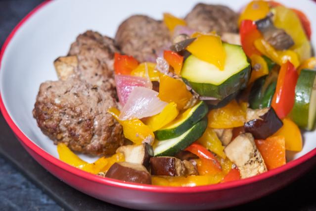 A bowl containing several browned meatballs served with colourful roasted vegetables. The vegetables include sliced courgette, red and yellow capsicum, red onion, and chunks of eggplant.