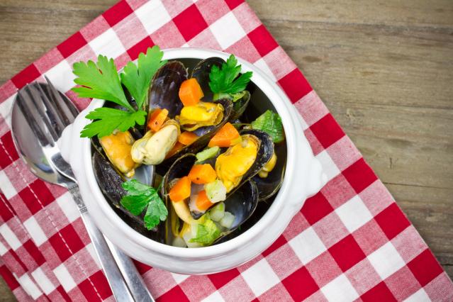 A white bowl of cooked mussels in their shells, mixed with diced carrot and chopped bok choy. The bowl is garnished with fresh parsley and sits on a red-and-white checkered cloth beside a fork and spoon on a wooden table.