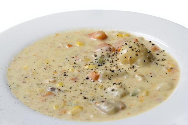 A white bowl filled with creamy mussel chowder. The chowder contains pieces of mussel, potato, carrot, celery, and sweetcorn in a thick pale broth, topped with a sprinkling of black pepper.