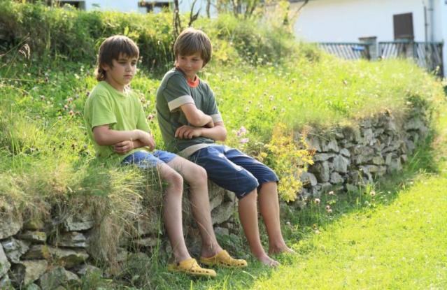 Two boys are sitting on a stone wall in a grassy area on a sunny day. One boy in a green shirt appears thoughtful or upset, while the other boy, in a gray shirt, looks at him with a slight smile, both with their arms crossed.