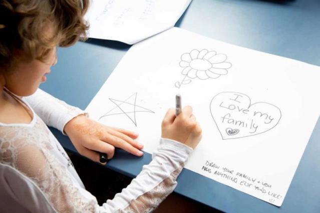 A child with wavy hair is seated at a table, drawing on a piece of paper. The paper features a star, a flower, and a heart with the words "I love my family" inside it. A prompt written on the paper reads, "Draw your family & you plus anything else you'd like!"