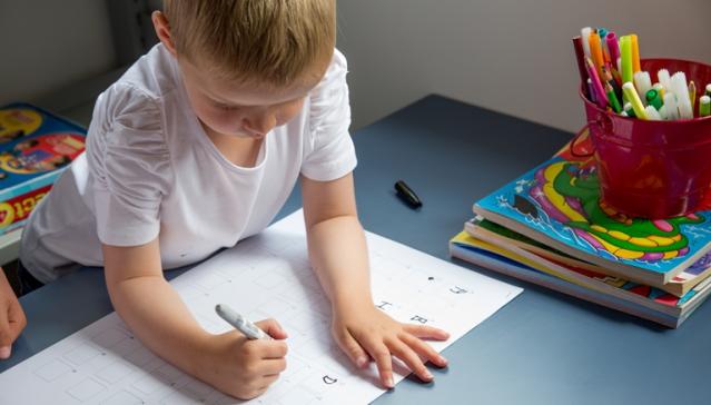 A young child is focused on drawing or writing with a marker on a worksheet at a table. Beside them is a red container filled with colorful markers and a stack of children's books.