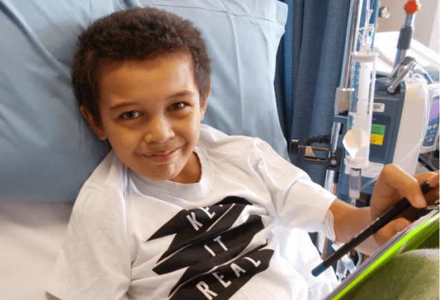 A young boy with short curly hair is lying in a hospital bed, smiling at the camera. He is holding a tablet and wearing a white T-shirt with the words "Keep It Real" printed on it. Medical equipment is visible in the background.