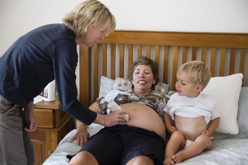 Lead maternity carer examining a woman lying on her bed during an antenatal check and woman's other child looks on