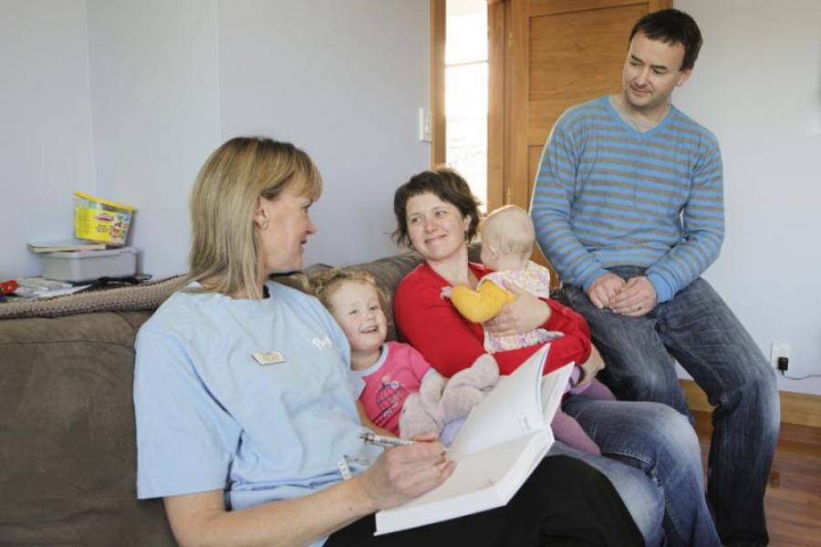 Well Child nurse sitting on a couch with a family (mother, father, baby and young child) during a home visit