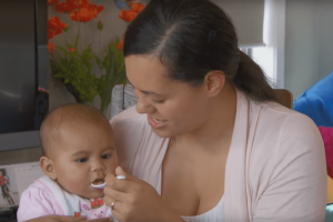 Mother giving her baby some solid food on a spoon while baby sits on her lap