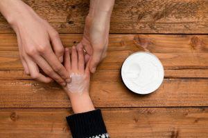 Photo of a woman's hands, putting cream on to a child's hand. Background is a table and a pottle of cream