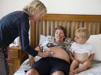 A pregnant mother lying on her bed. Her young child is looking on as the lead maternity carer examines the mother's tummy